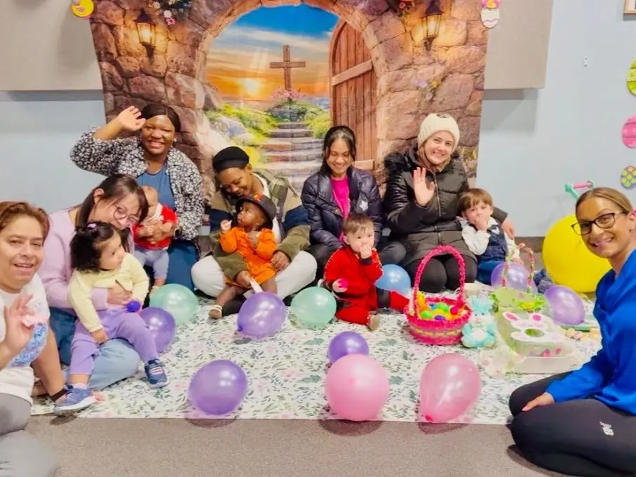 Seven adults and five babies sit on a mat decorated with balloons and Easter theme