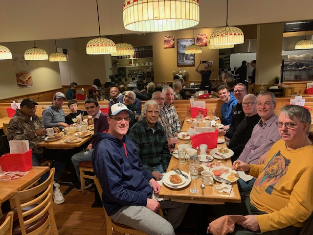 Fifteen men sit at two tables over breakfast